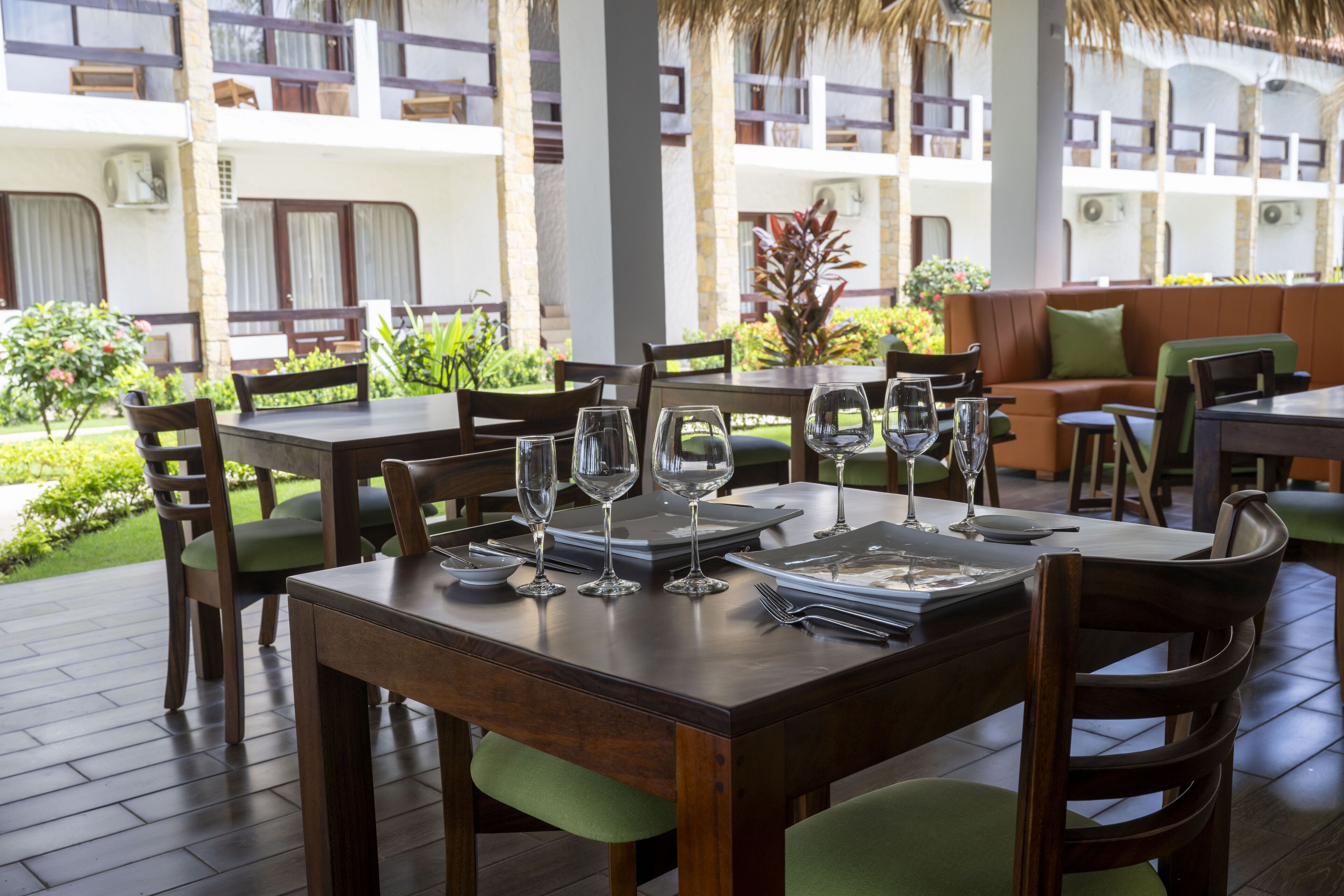Un comedor con mesas y sillas de madera, techo de paja y vista a una piscina y un edificio al fondo.