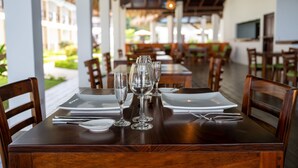 A dining table set with plates, cutlery, and wine glasses, surrounded by wooden chairs in a restaurant with a view of greenery. - Fuego del Sol Beachfront Hotel (Jacó)