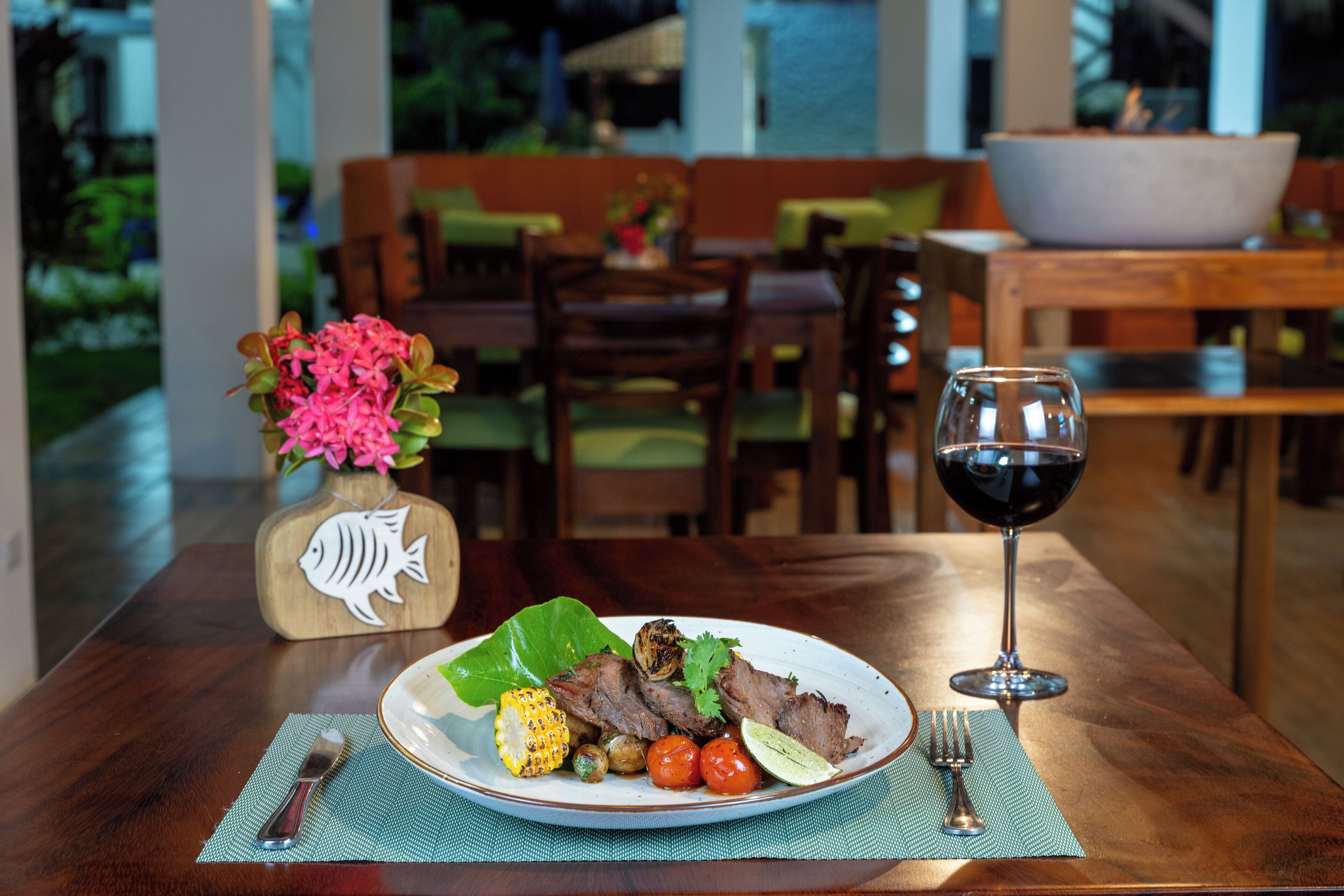 A dining table with a plate of food, a glass of red wine, and a decorative fish-shaped vase.