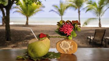 A coconut drink with a straw and a strawberry on a table with a wooden fish decoration, set against a beach view with palm trees.