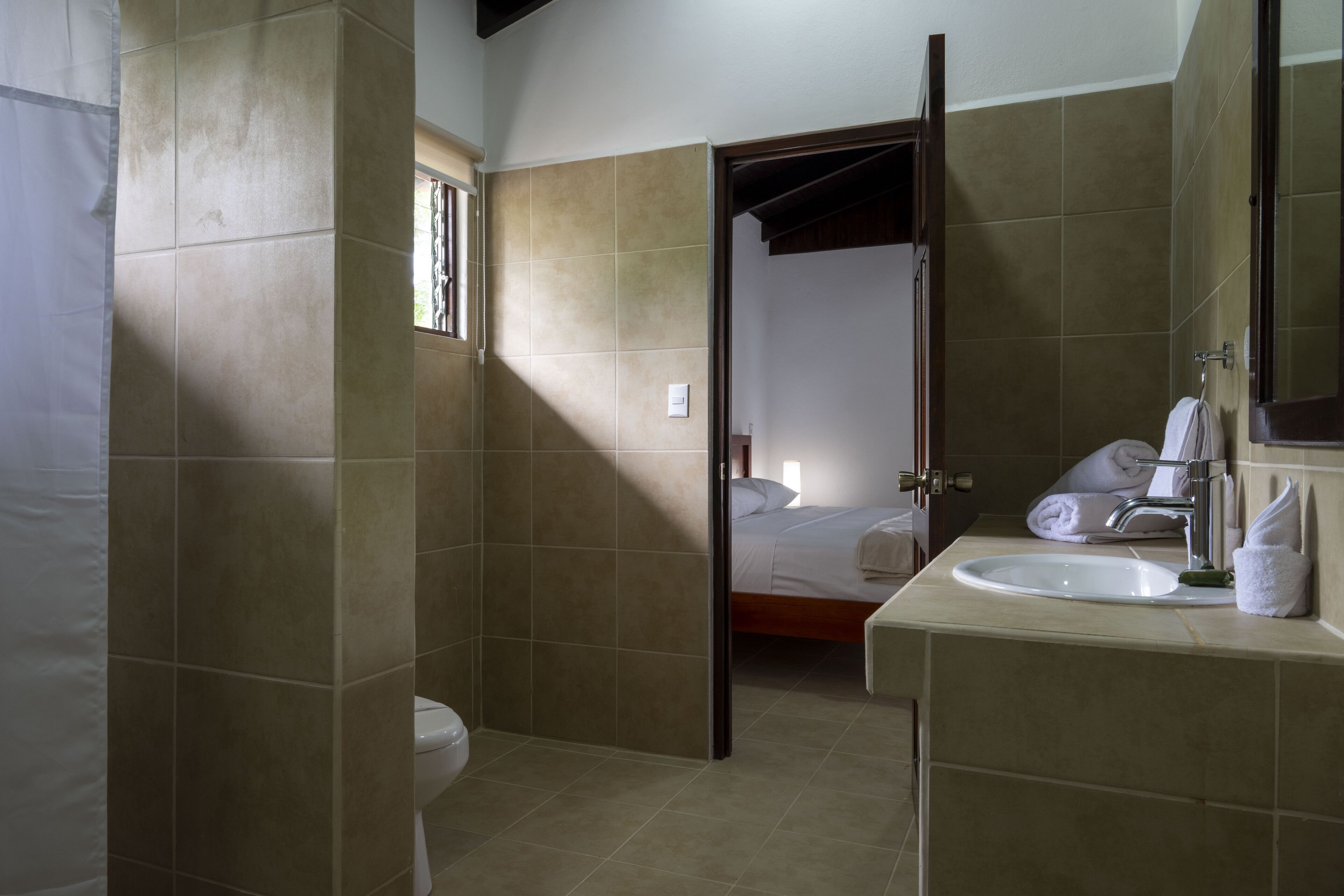Een badkamer met een wastafel, toilet en douchegedeelte. In de verte is een bed te zien door een geopende deur.