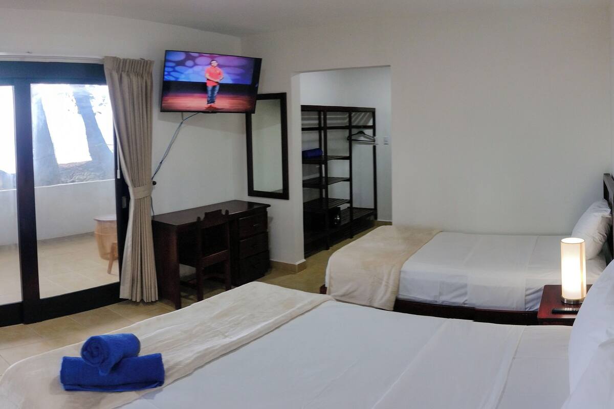 A hotel room with two beds, a TV on the wall, a desk with a chair, a wardrobe, and a window with curtains.