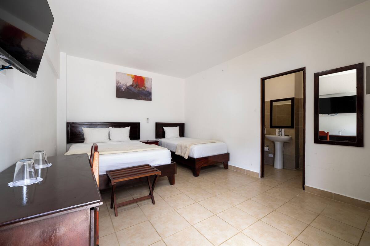 A hotel room with two beds, a desk, a TV, a painting, a mirror, and a bathroom.