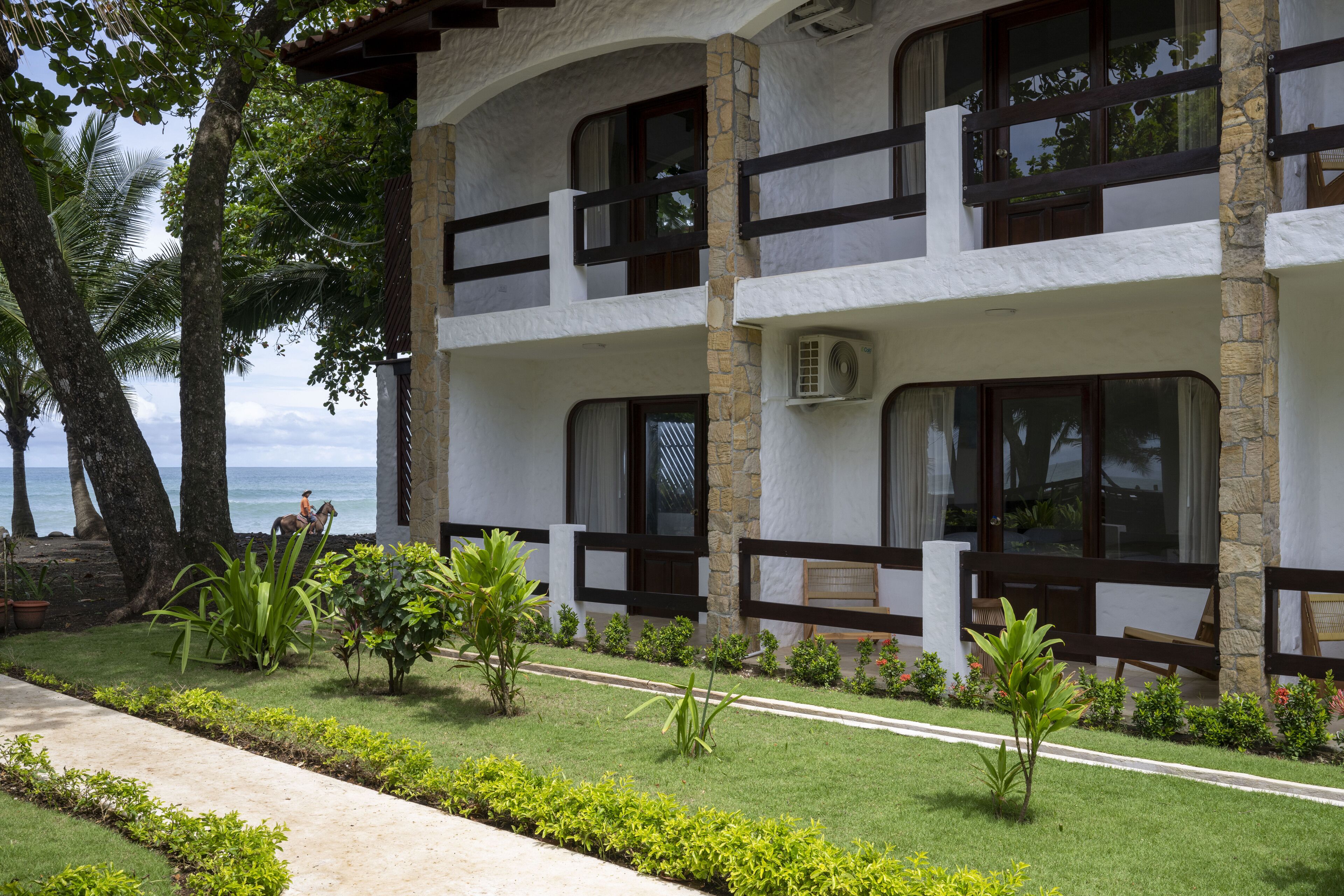 Un edificio de dos pisos con un balcón, rodeado de vegetación tropical y vistas al océano.