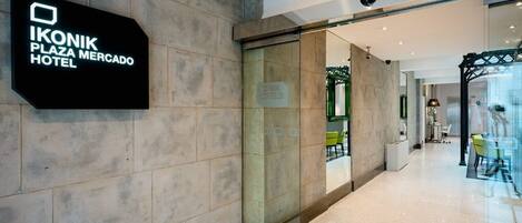 A modern hotel entrance with a glass door, a sign for the Ikonik Plaza Mercado Hotel, and a clean, tiled floor.