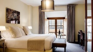 A hotel room with a large bed, bedside lamps, a bench, a small table with a bottle, and a view of the outside through a window. - Ikonik Plaza Mercado (Salamanca)
