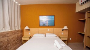 Una habitación de hotel con cabecera de madera, una cama grande con sábanas blancas, dos mesitas de noche con lámparas, y un cuadro de un paisaje en la pared.