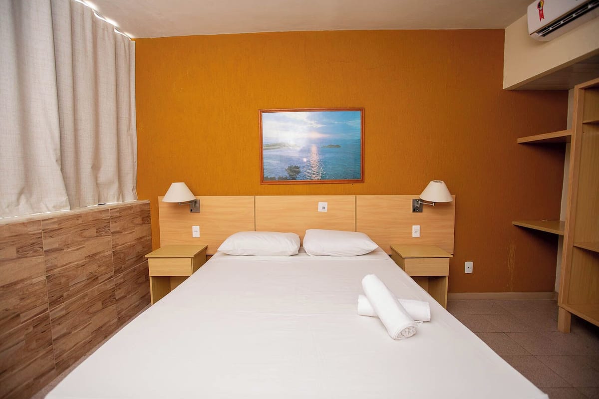 A hotel room with a wooden headboard, a large bed with white linens, two bedside tables with lamps, and a painting of a landscape on the wall.