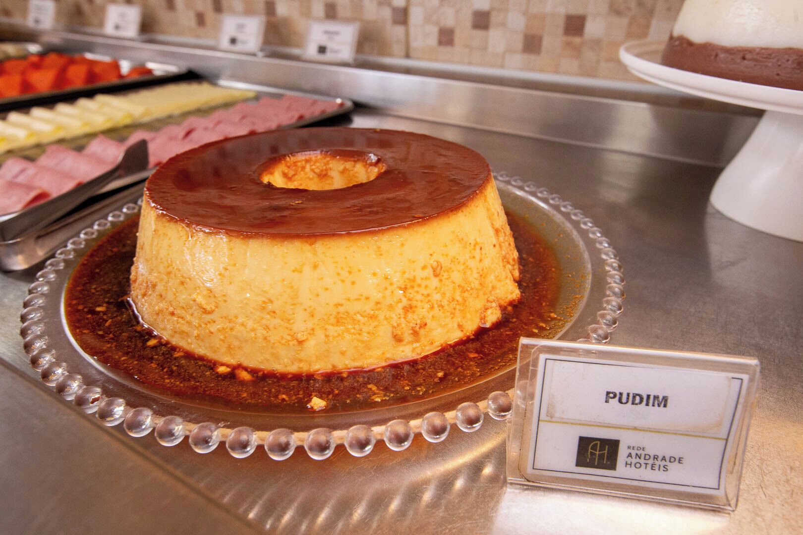 En dessert med teksten 'PUDIM' på en metalserveringsbakke med et navneskilt.