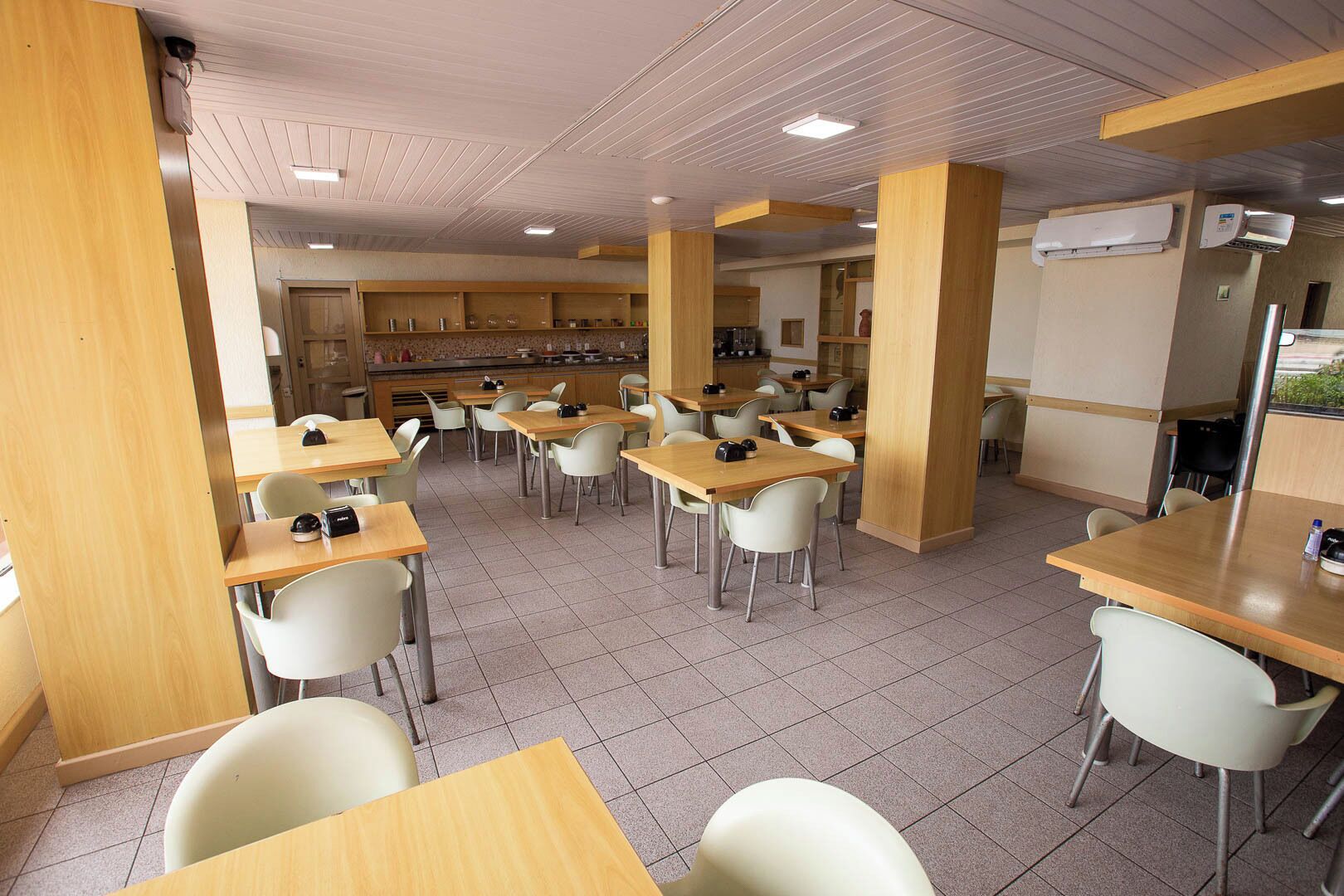 A cafeteria with multiple tables and chairs, wooden pillars, and tiled flooring.