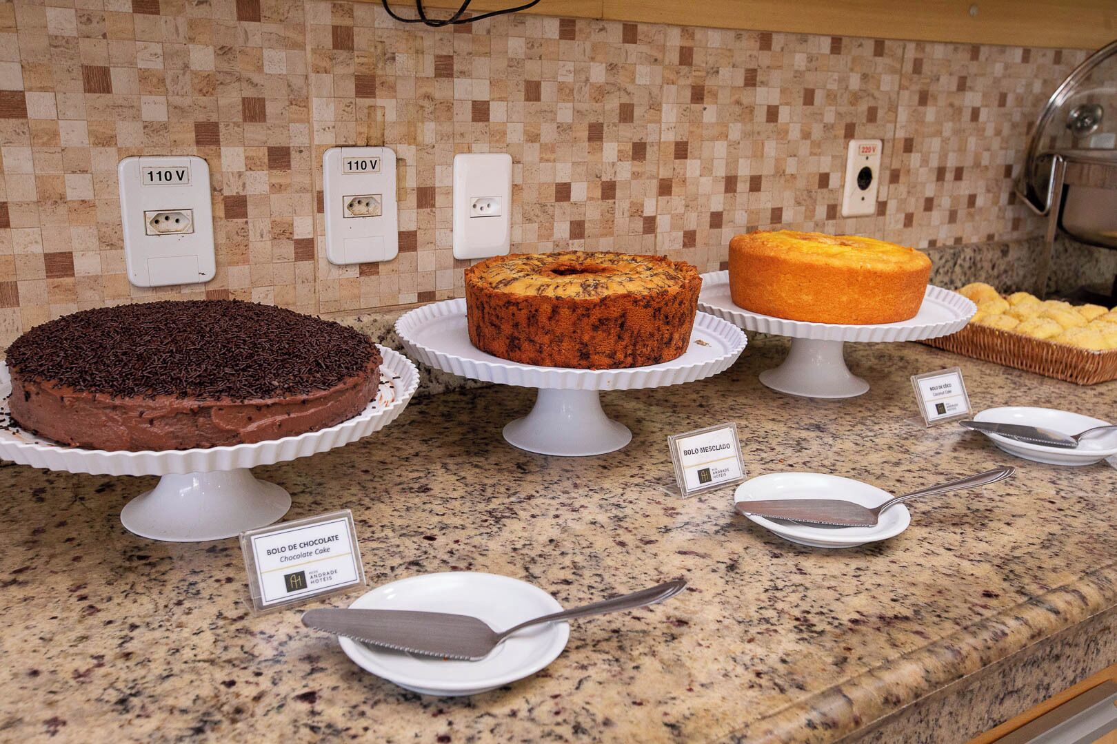 A kitchen counter with three cakes on white stands, each labeled with a different name.
