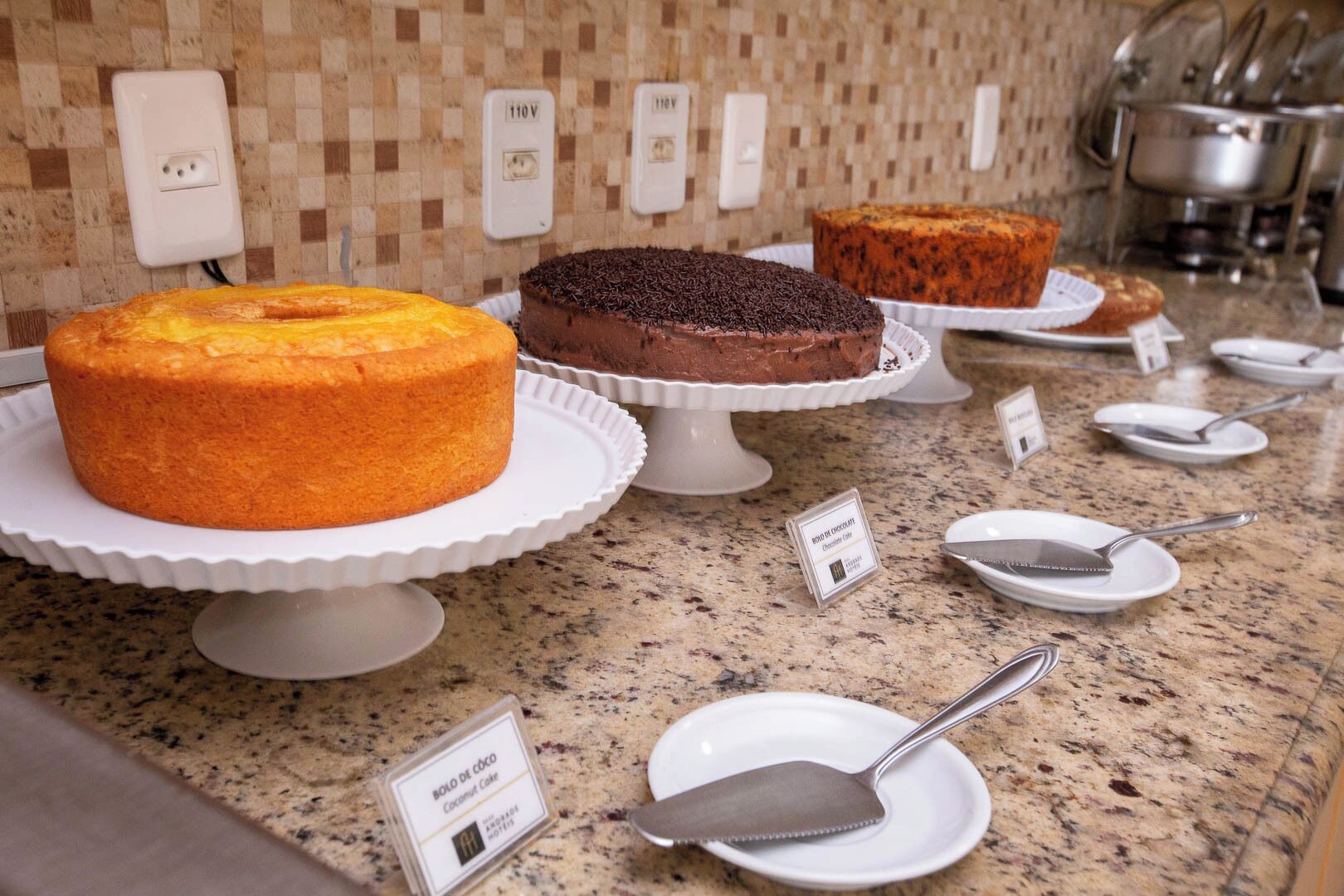 A buffet counter with various cakes on white stands, a granite countertop, and a tiled backsplash with electrical outlets.