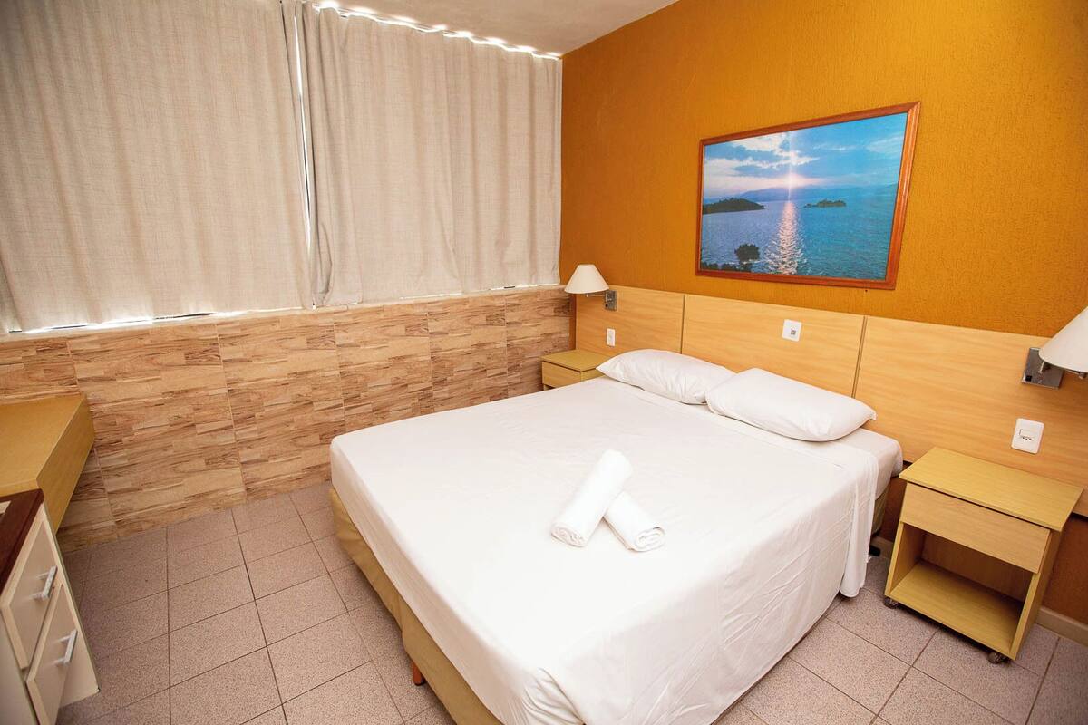 A hotel room with a large bed, a nightstand, a wooden headboard, a painting of a lake, and a window with curtains.