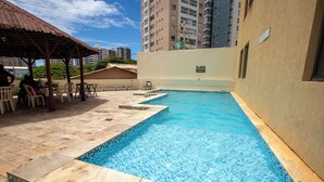 Una piscina con bordes de azulejos, un área de descanso cubierta con sillas, y un edificio al fondo.