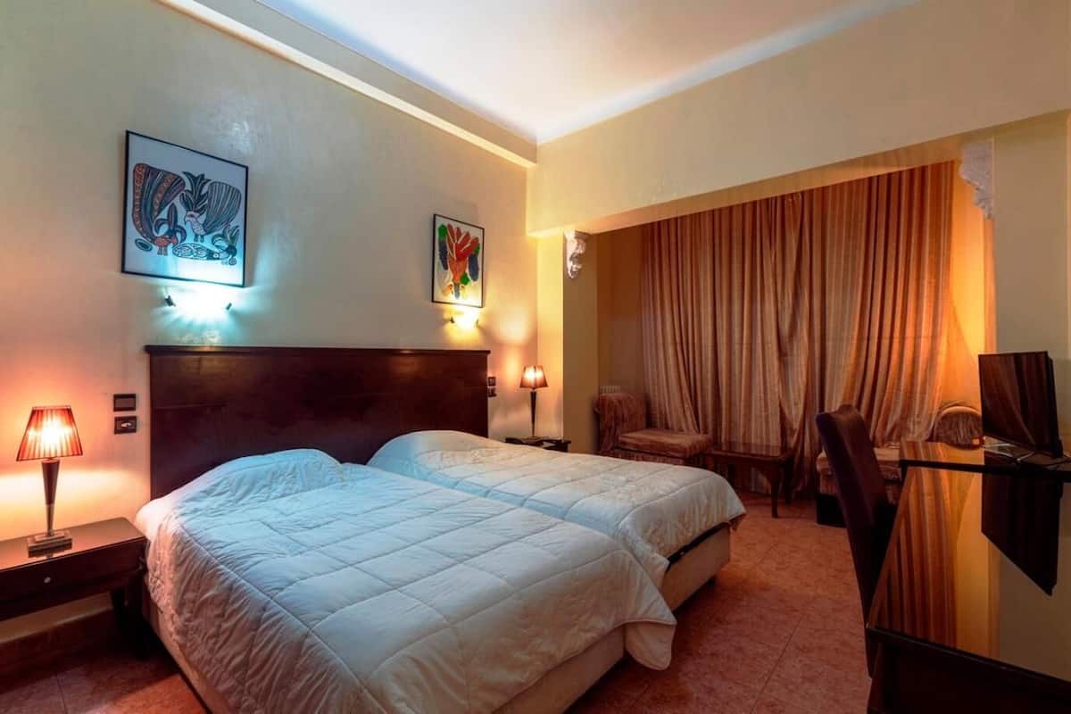 Een hotelkamer met een groot bed, twee nachtkastjes met lampen, een stoel, een bureau en een groot raam met gordijnen.