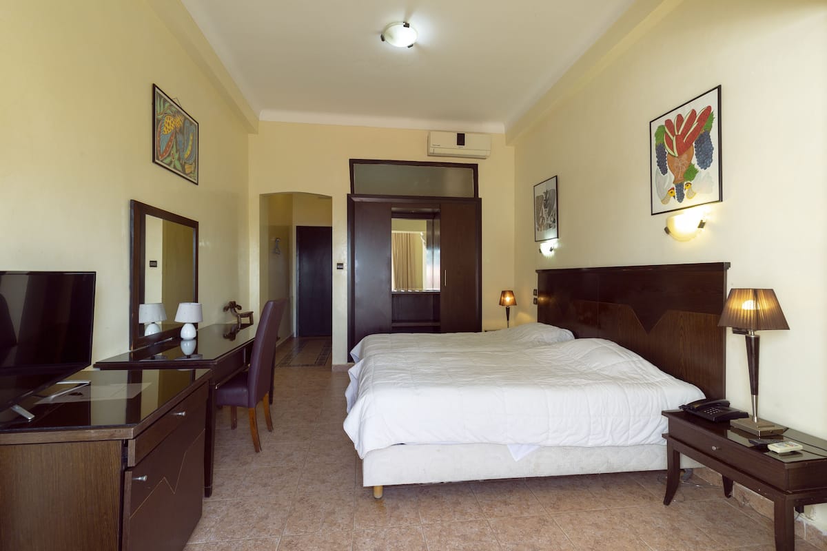 Een hotelkamer met een bed, een bureau met een televisie, een stoel, een nachtkastje met een lamp, en kunstwerken aan de muur.