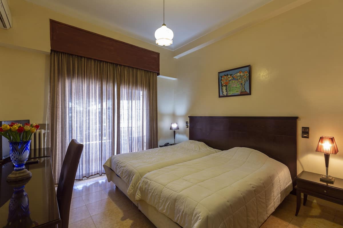 Een hotelkamer met een groot bed, een nachtkastje met een lamp, een stoel, een bureau met een bloevas en een raam met gordijnen.
