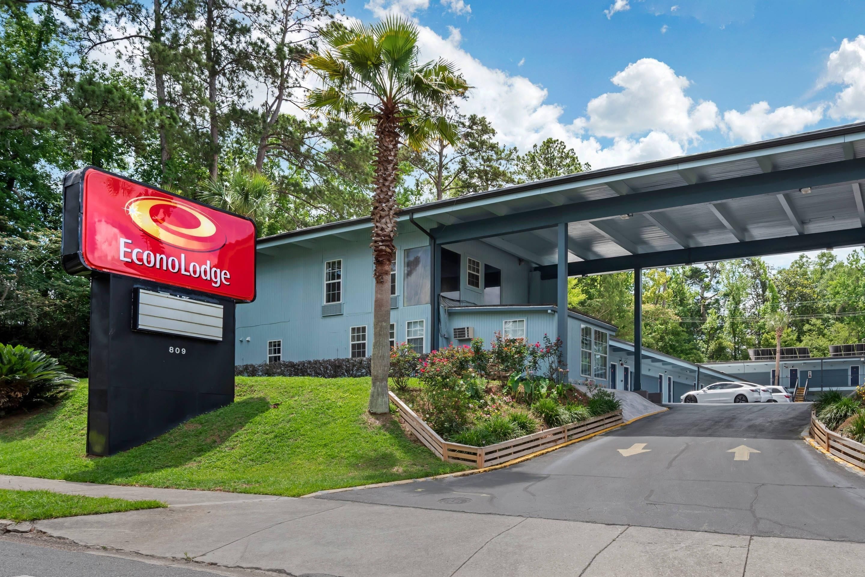 A hotel entrance with a red sign, a covered walkway, and a parking area.