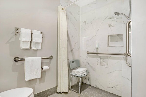 A bathroom with a shower, a toilet, a grab bar, a chair, and towels. - Econo Lodge Tallahassee Downtown - University (Tallahassee)