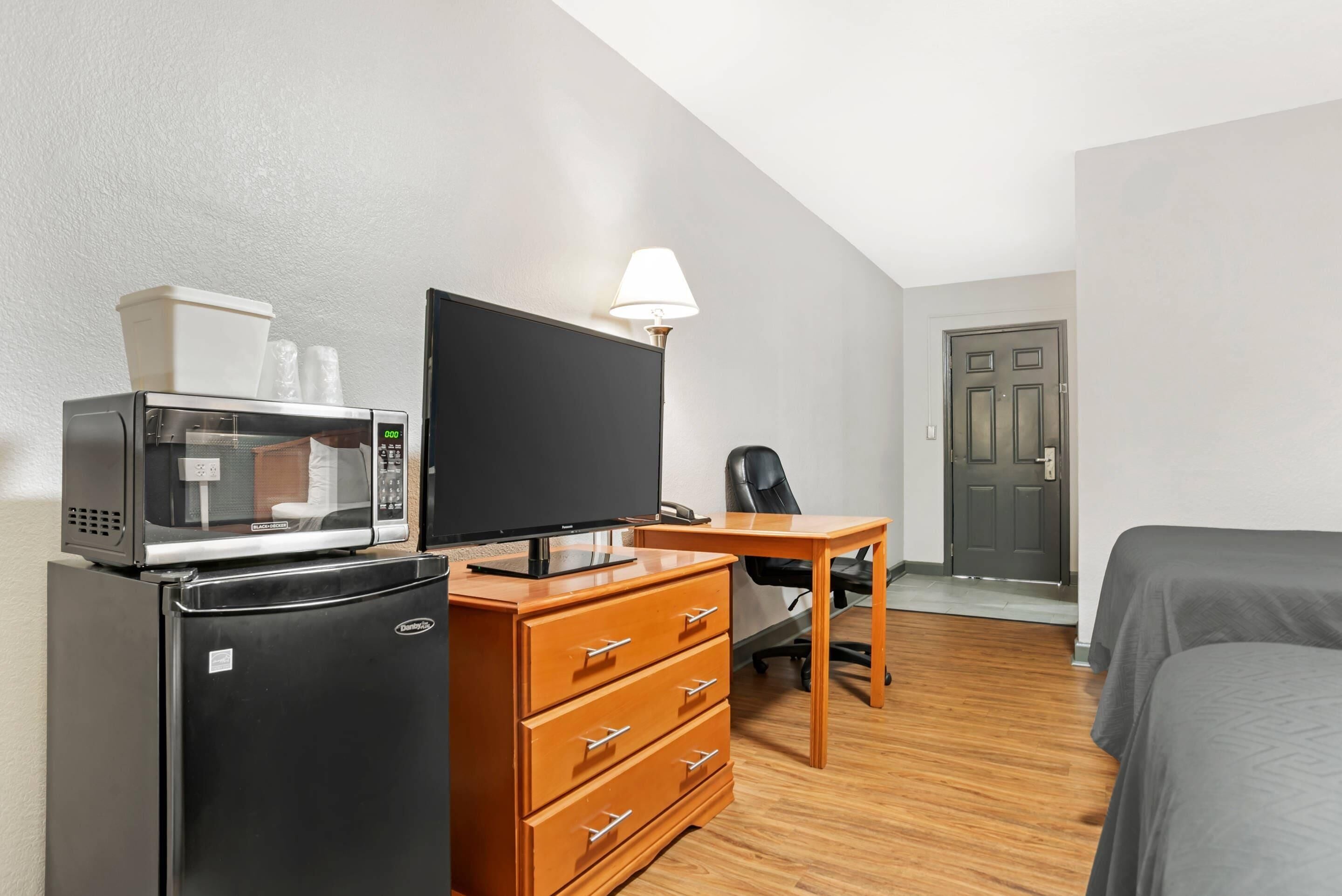 A compact hotel room with a kitchenette, a flat-screen TV, a wooden desk, a chair, and a bed.