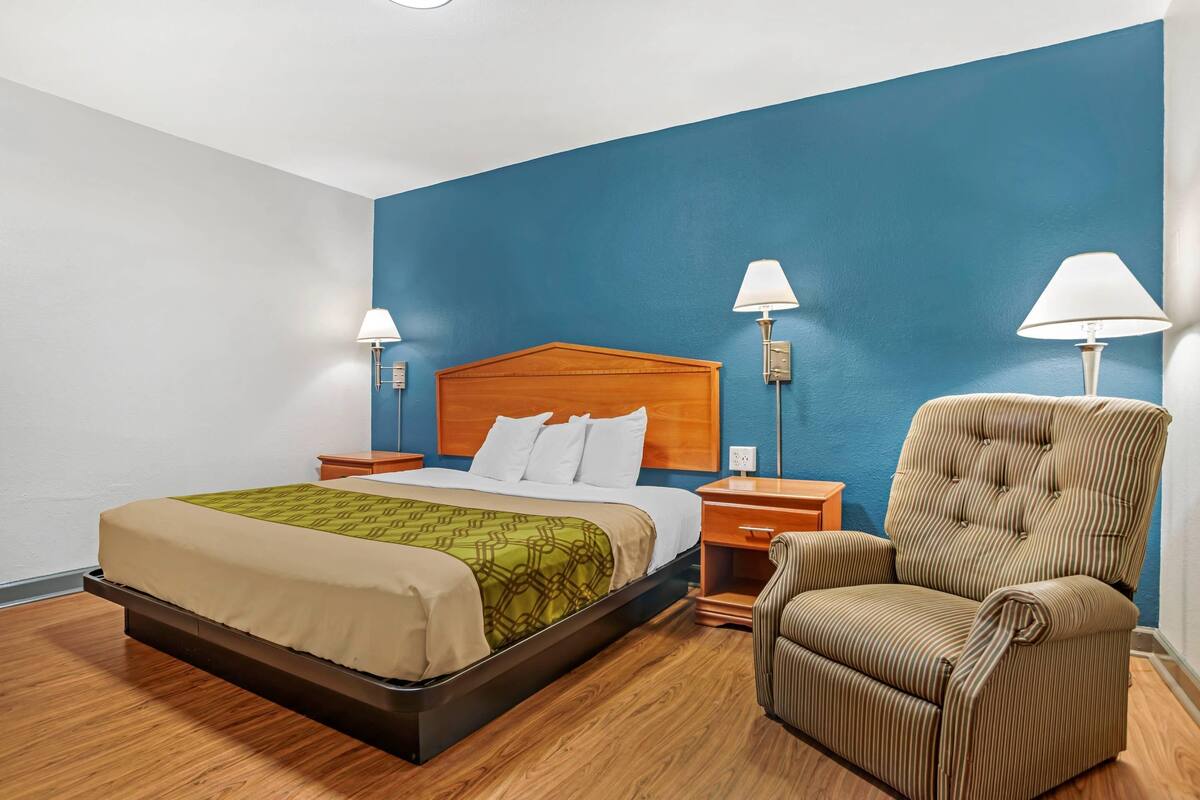 A hotel room with a bed, two bedside lamps, a chair, and a wooden headboard.