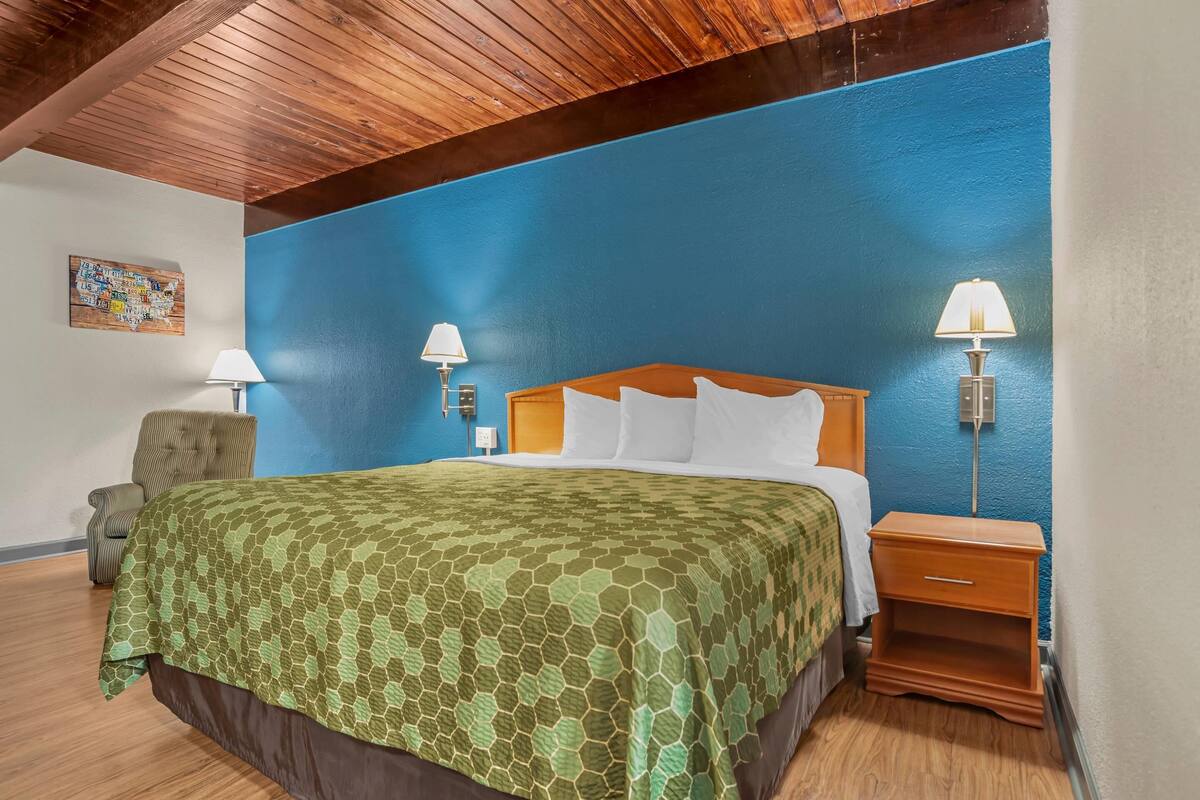 A hotel room with a wooden ceiling, a large bed with a green patterned bedspread, two bedside lamps, a wooden nightstand, and a chair.