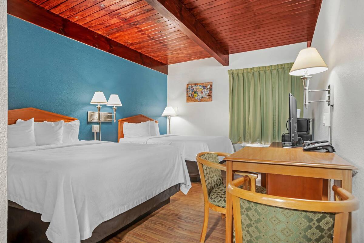 A hotel room with two beds, a wooden desk, chairs, lamps, a television, and a green curtain.