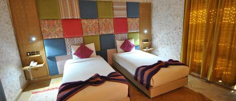 A hotel room with two beds, a decorative headboard, bedside tables with lamps, a wooden floor, and a window with curtains.