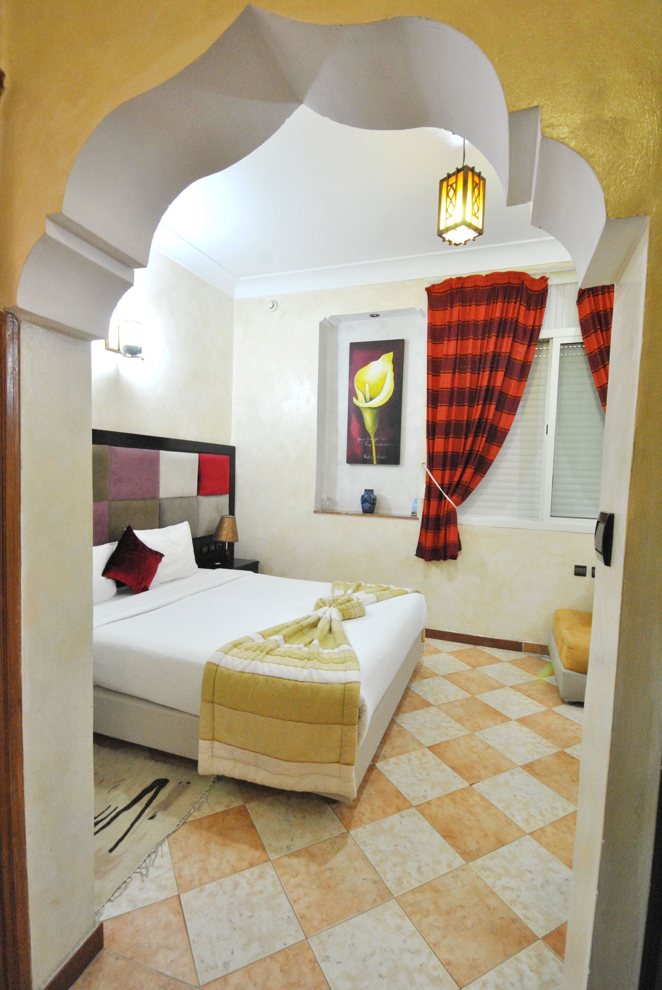 Quarto com cama, mesa de cabeceira, abajur, janela com cortinas e um vaso fixado na parede.