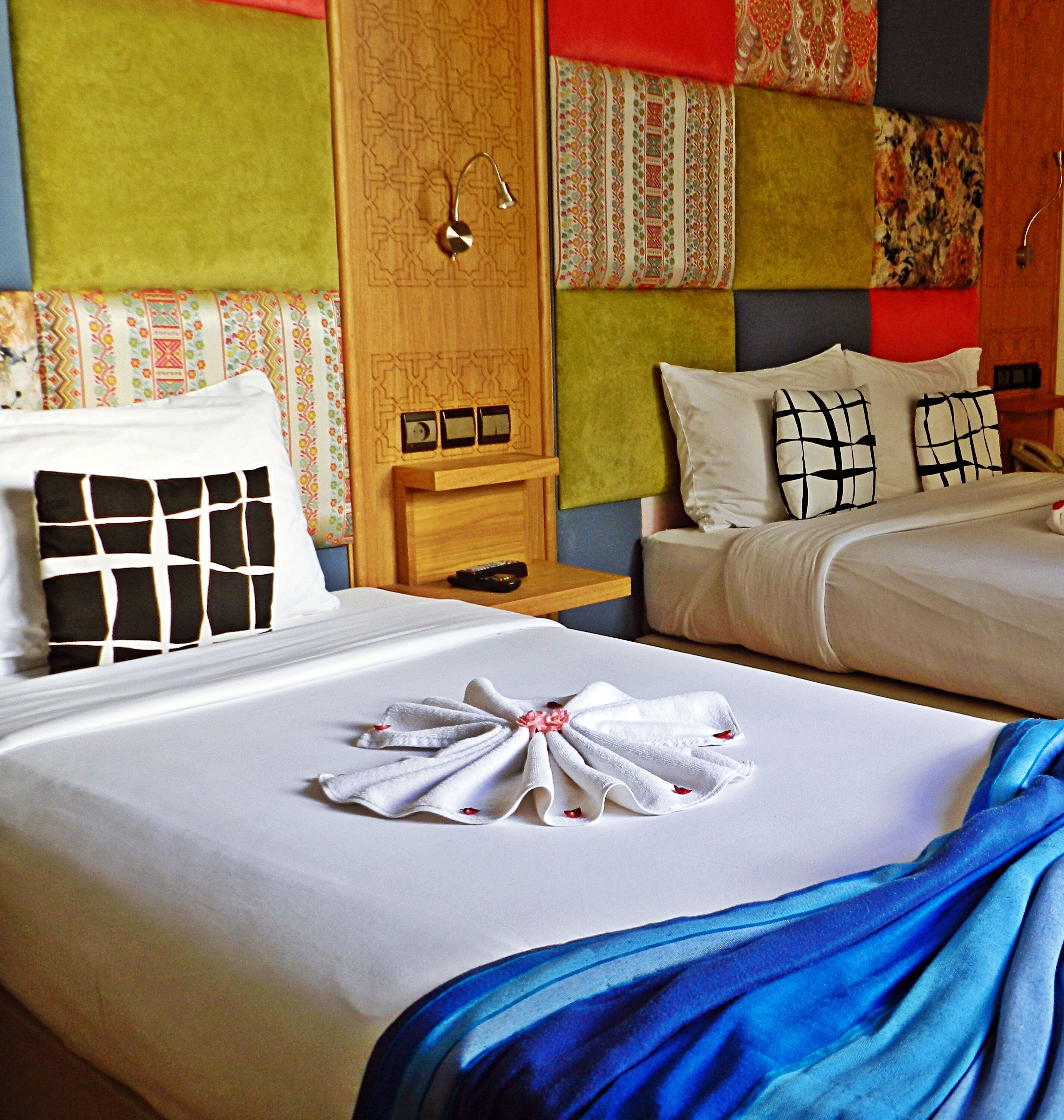 Habitación de hotel con dos camas, cabecero de madera, mesita de noche con lámpara y pared con motivos coloridos.