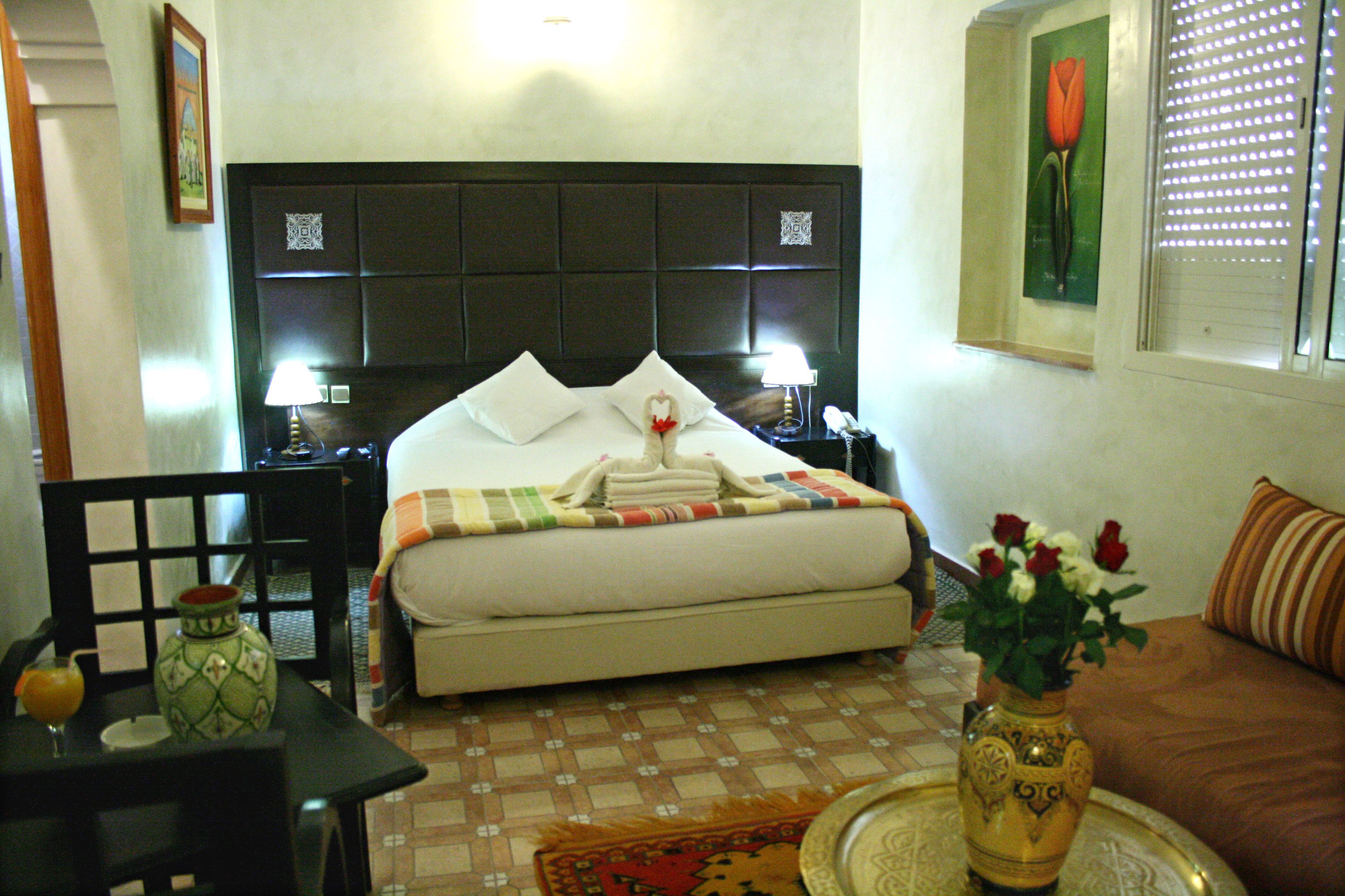 A hotel room with a bed, bedside tables, a vase with flowers, a chair, and a dining table.