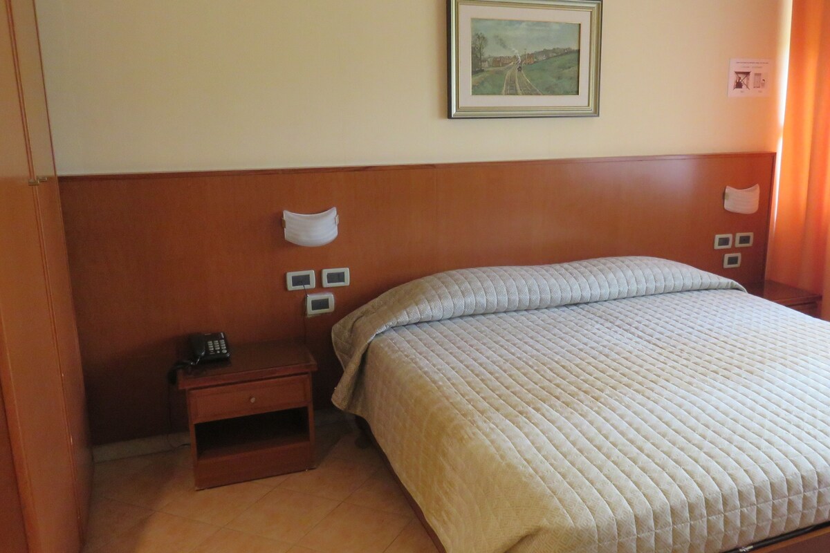 A hotel room with a bed, a nightstand, a painting on the wall, and a window with curtains.