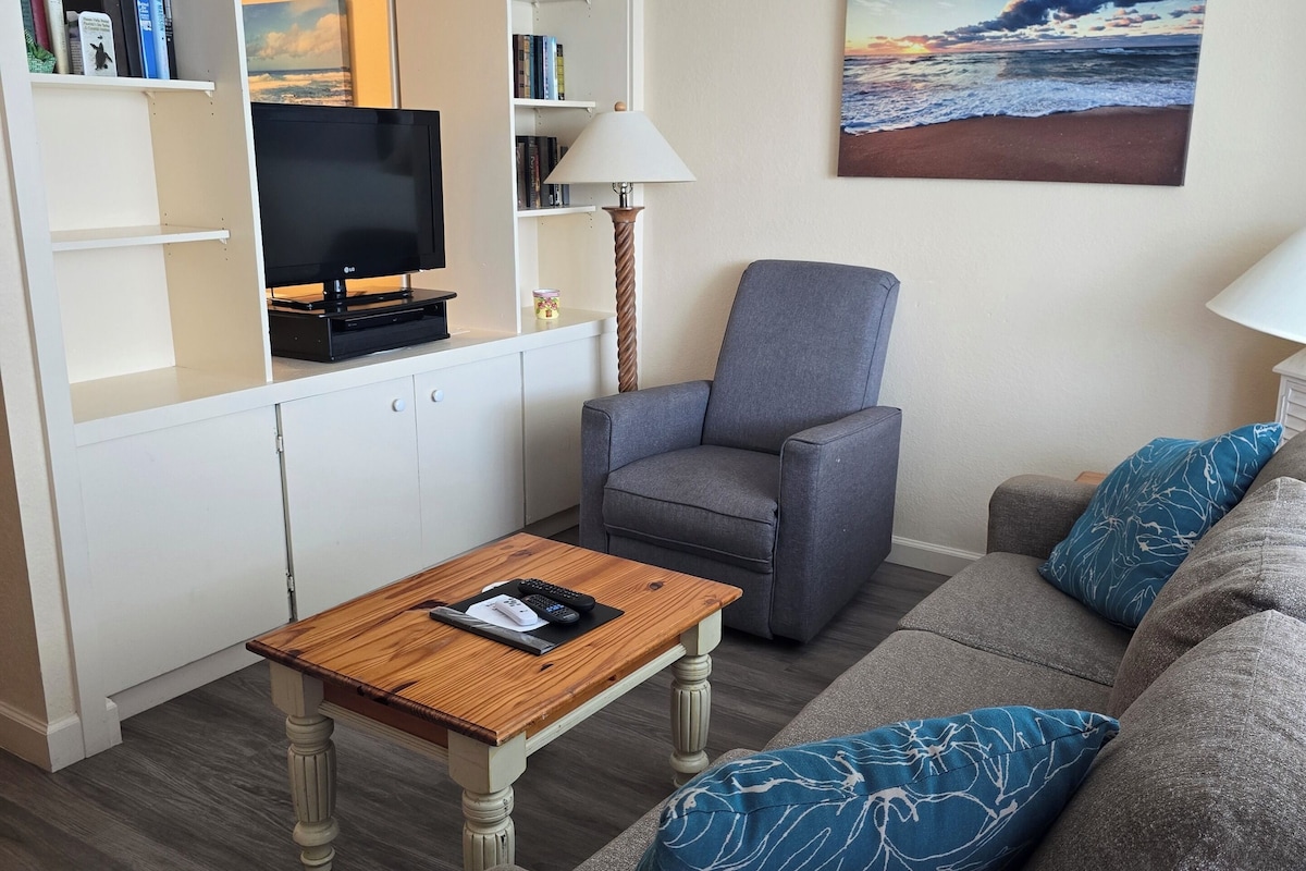 Een woonkamer met een grijze bank, een houten salontafel, een televisie en een boekenkast.