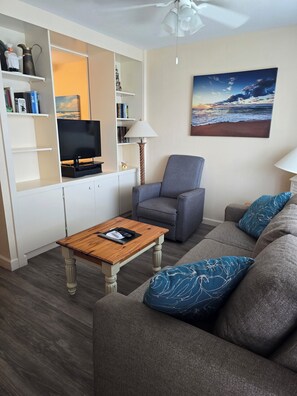 A living room with a grey sofa, a wooden coffee table, a television, and a bookshelf. - The Driftwood Resort (Vero Beach)