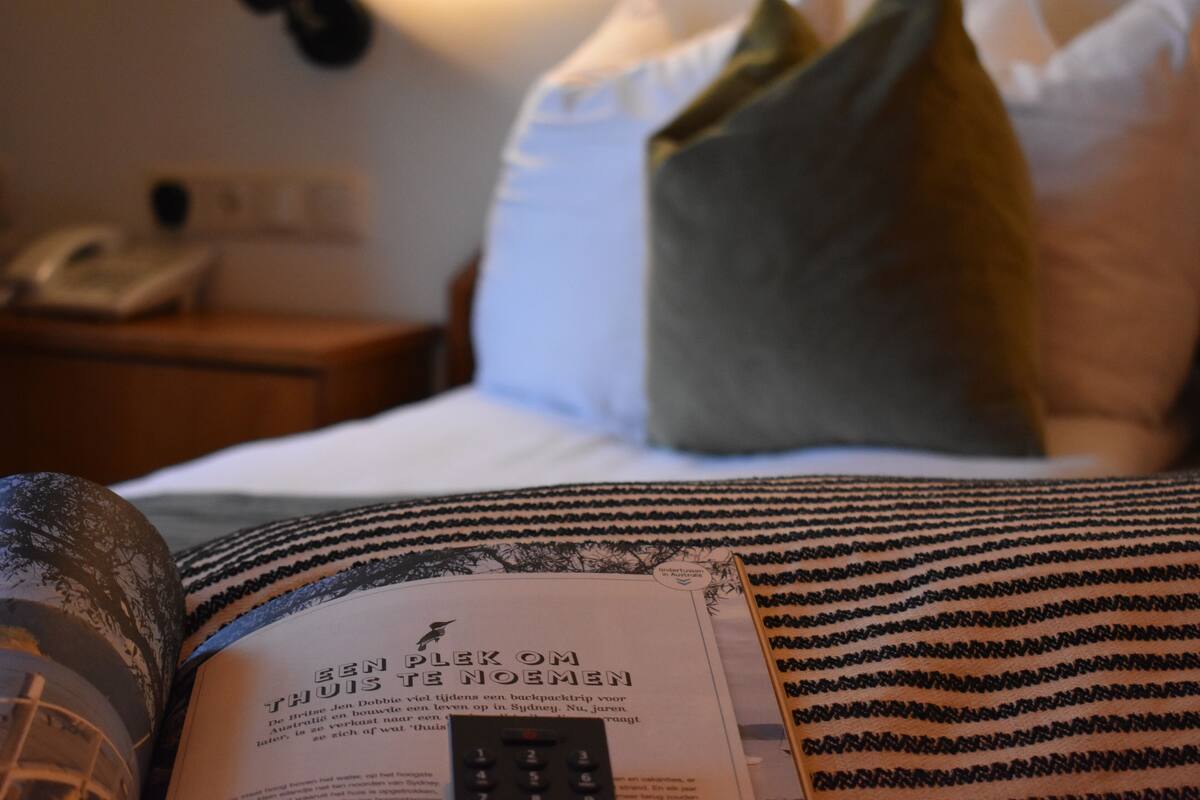 Una habitación de hotel con cama, almohadas, un control remoto y una revista sobre la cama.