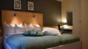 Una cama impecablemente hecha con una colcha verde y blanca, varios almohadones y un cabecero de madera. Se observa un mesita de noche con una lámpara y un teléfono, y un espejo en la pared.
