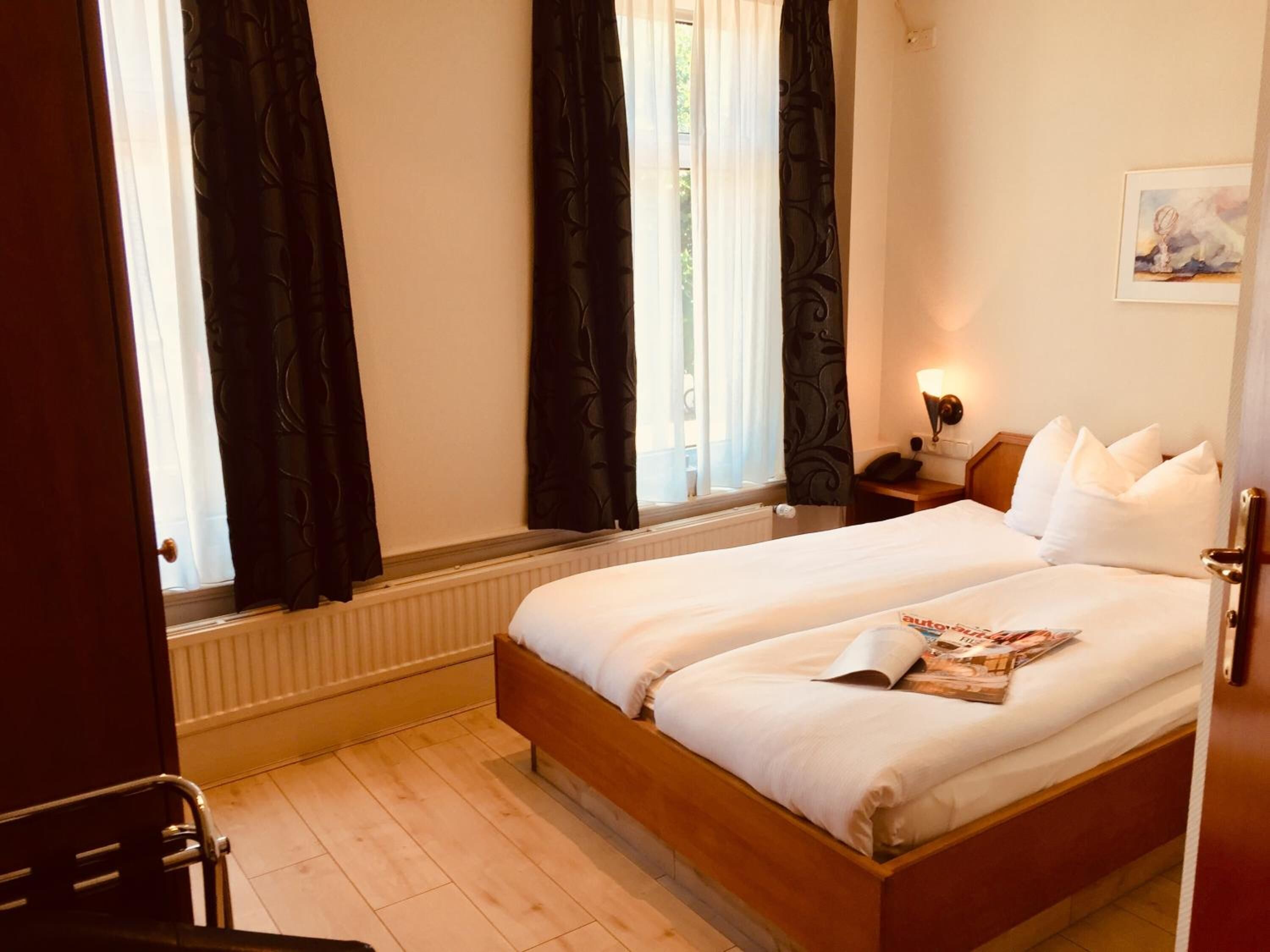 Een hotelkamer met een tweepersoonsbed, een nachtkastje met een lamp, een raam met gordijnen en een schilderij aan de muur.