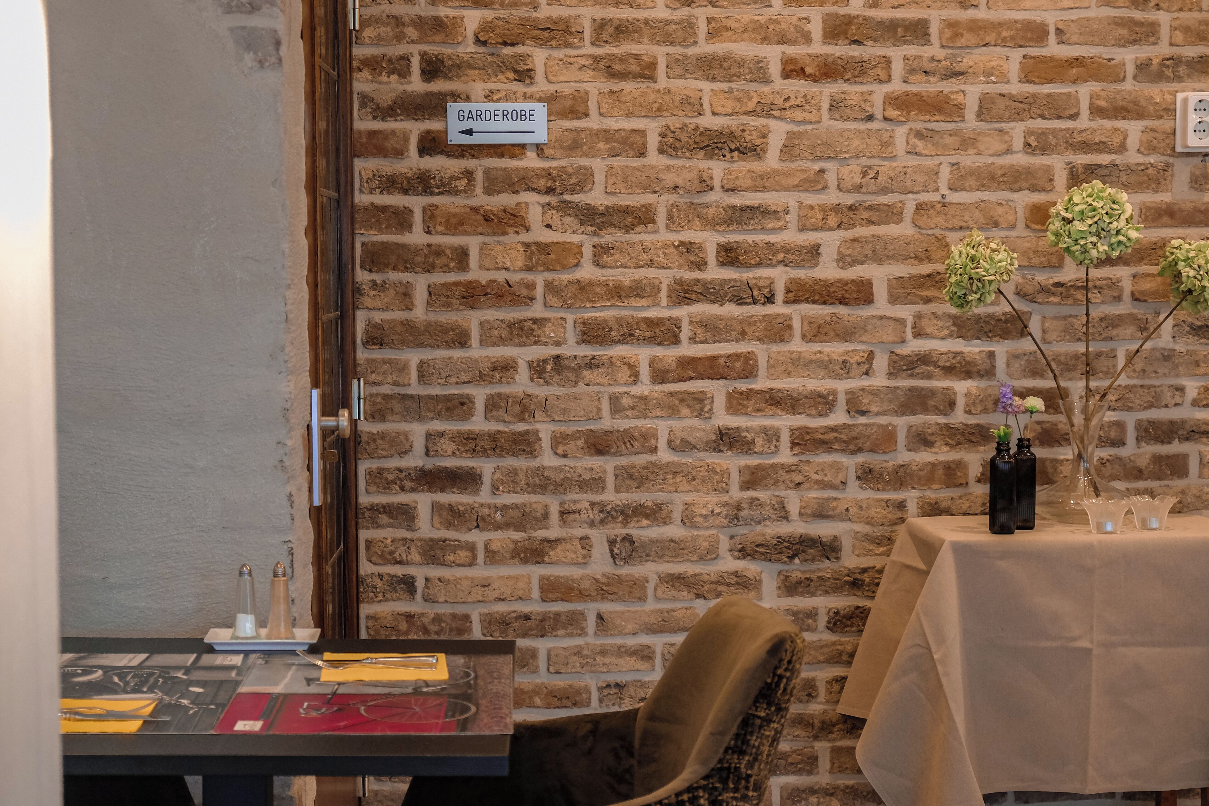 Een bakstenen muur met een bordje dat naar 'Garderobe' wijst, een tafel met een vaas en kaarsen, en een stoel.