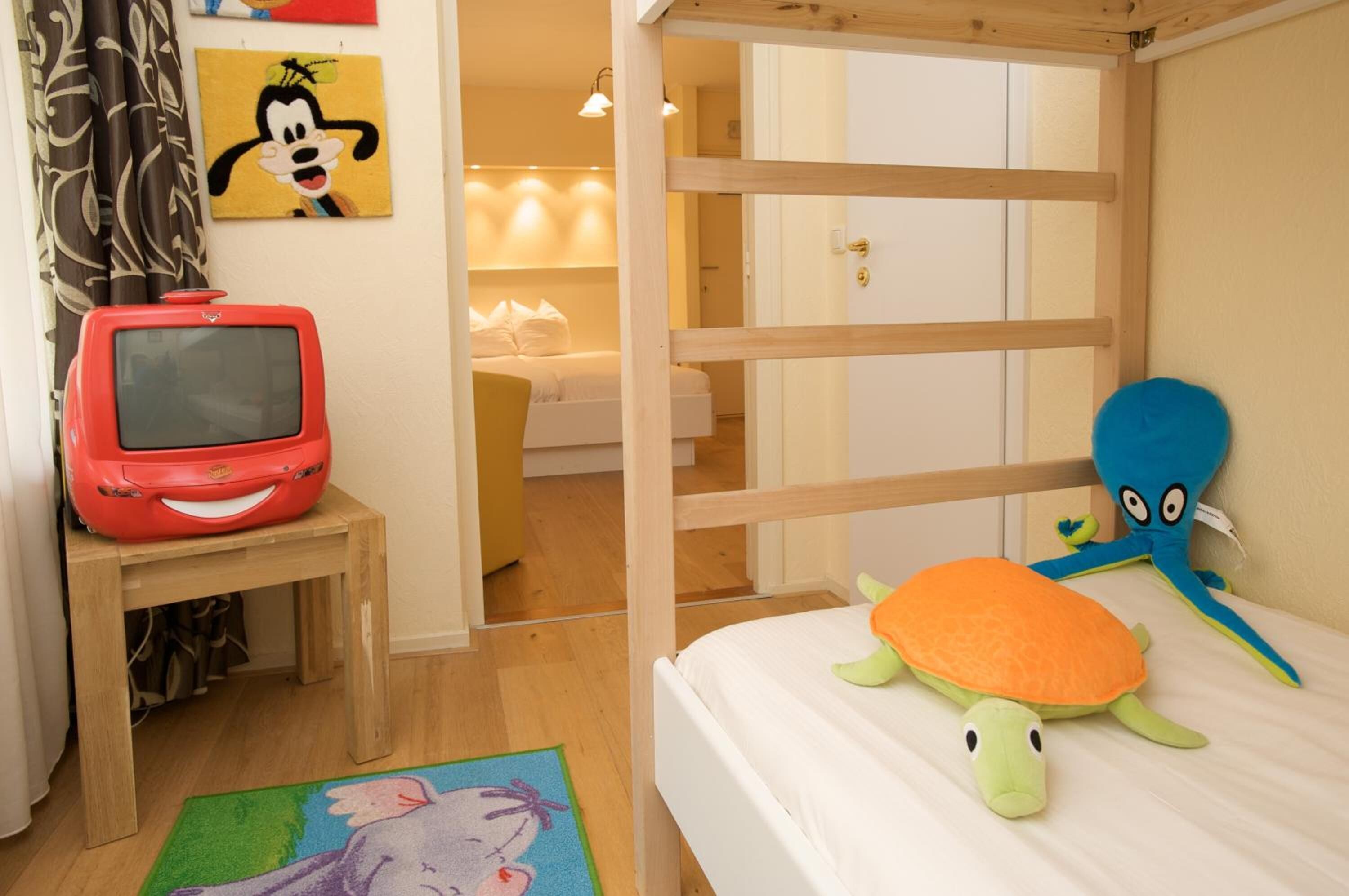 Quarto de criança com uma cama beliche, uma mesinha com uma televisão vermelha e brinquedos macios na cama.