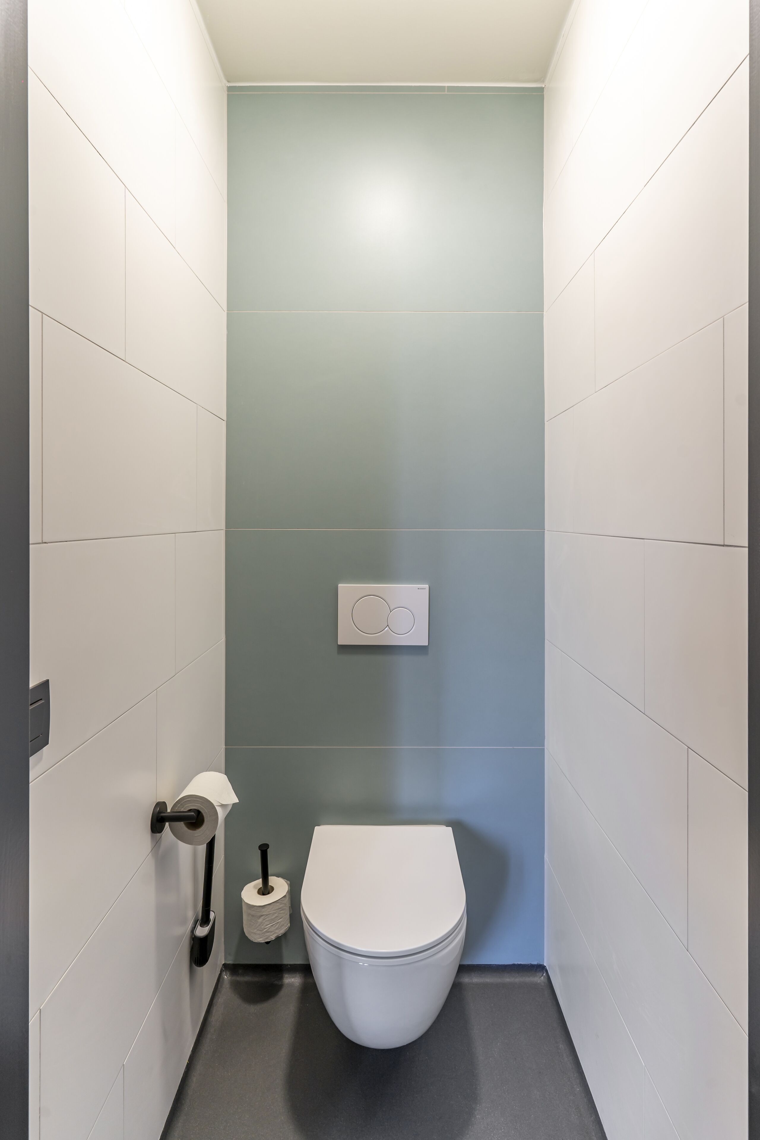 A small, modern bathroom with white and light blue tiles, a white toilet, a toilet paper holder, and a flush handle.