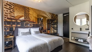 A modern bedroom with a brick-patterned headboard, a large bed, a bedside table with a lamp, and a round mirror above the dresser. - Dormio Wijnhotel Valkenburg (Valkenburg aan de Geul)