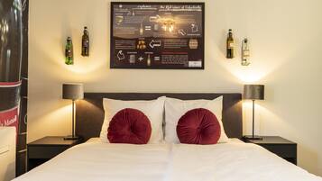 Habitación de hotel con un cartel informativo grande en la pared, dos mesitas de noche con lámparas, una cama con ropa blanca y almohadas rojas, y una mesita de noche con lámpara.