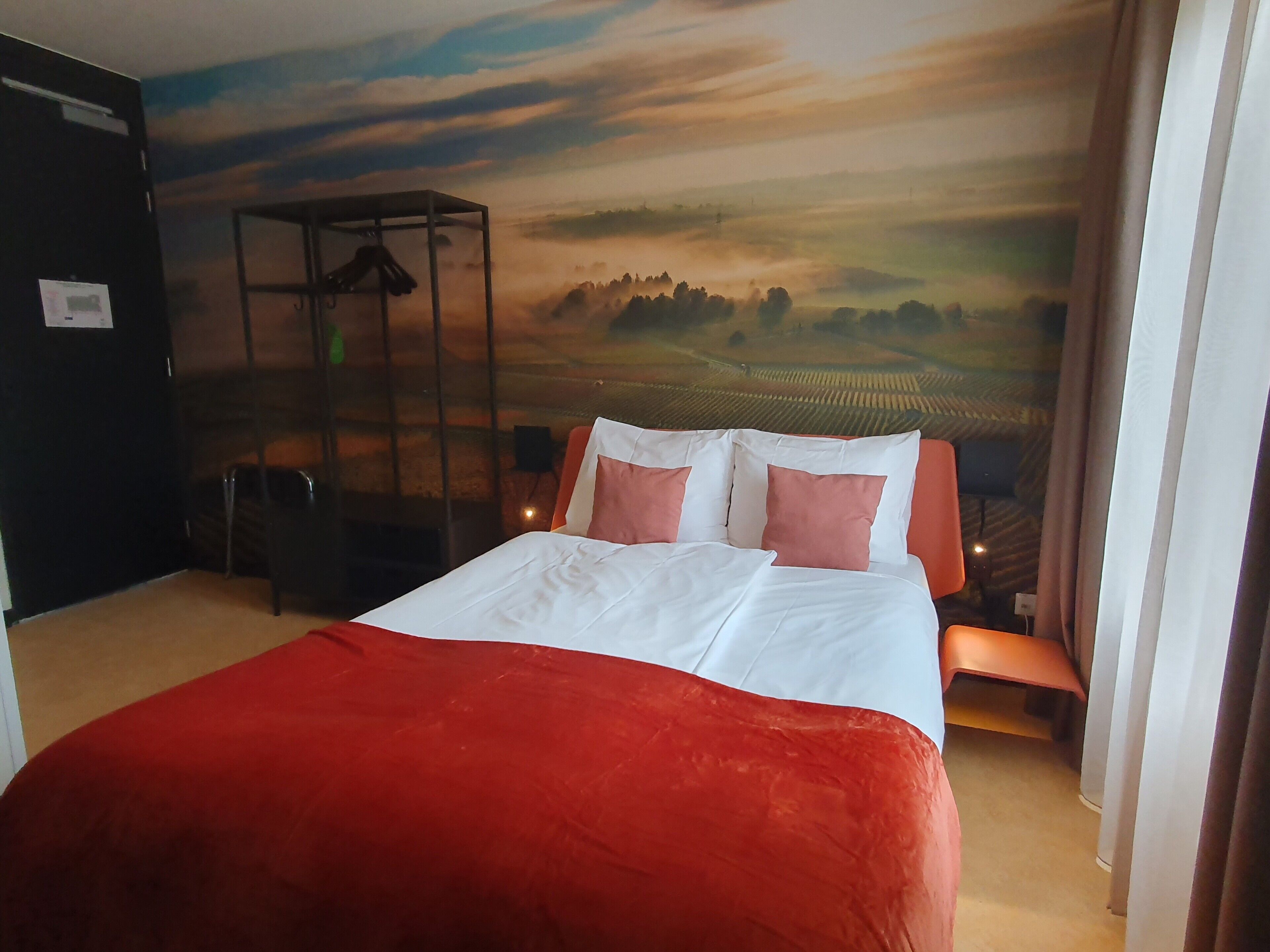 Une chambre d’hôtel avec un lit, une couverture rouge et une fresque représentant un paysage sur le mur.