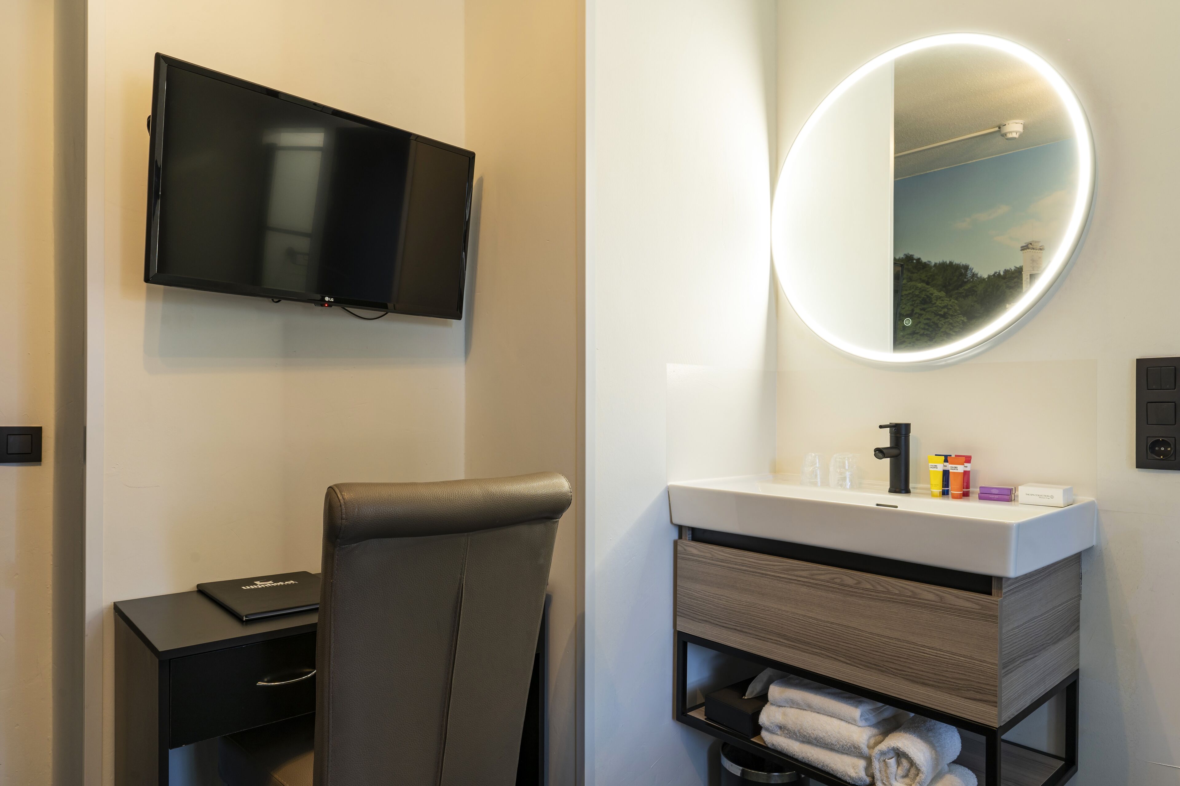 Et moderne hotelværelse med et fladskærms-tv på væggen, et rundt spejl med lys, en sort læderstol, et lille skrivebord med en bærbar computer og et sminkebord med toiletartikler.