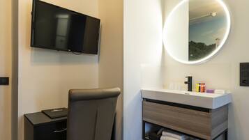 A modern hotel room with a flat-screen TV mounted on the wall, a round mirror with a light, a black leather chair, a small desk with a laptop, and a vanity with toiletries.