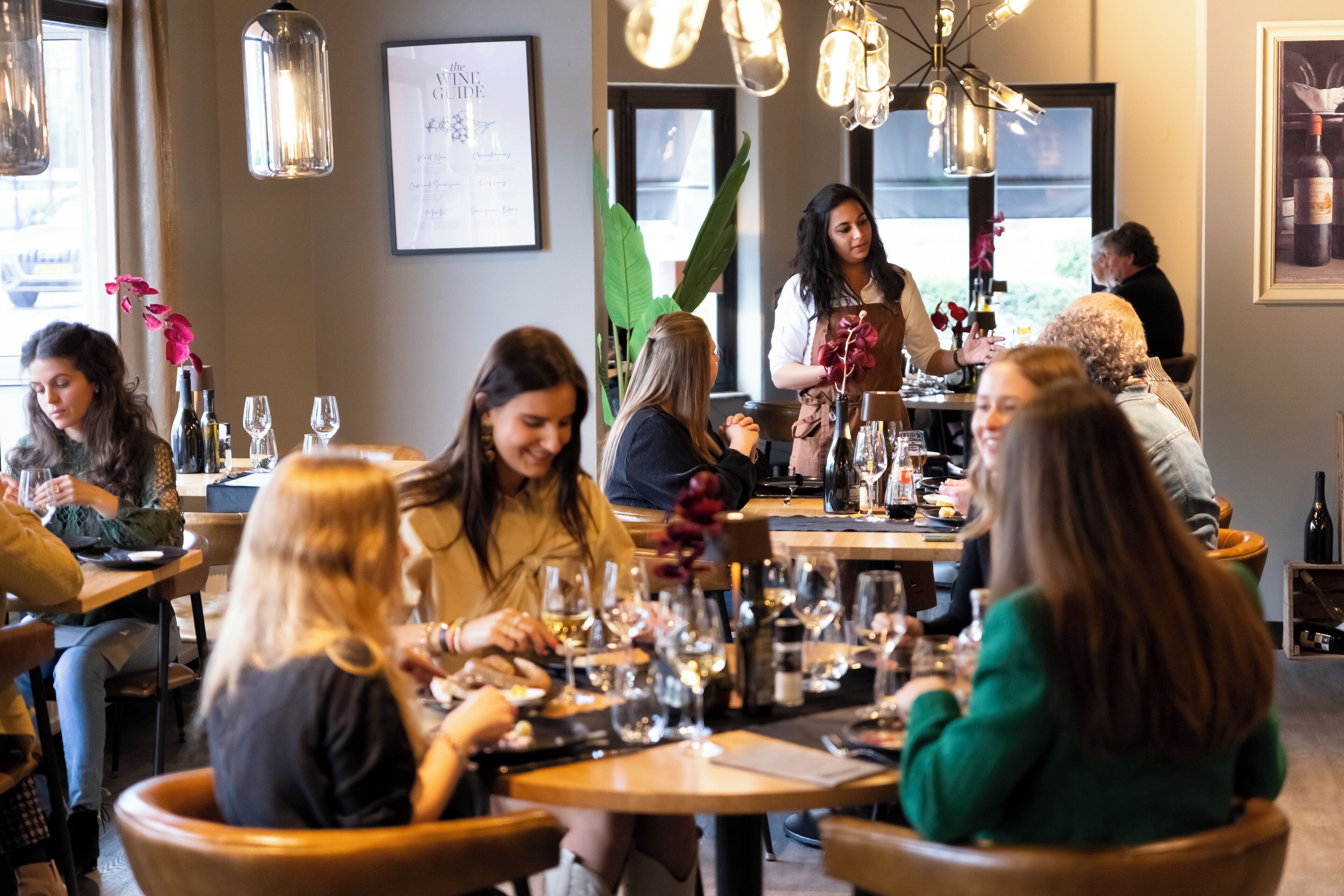 En restauranginteriör med runda bord, vinglas och en servitör som tar hand om gästerna.