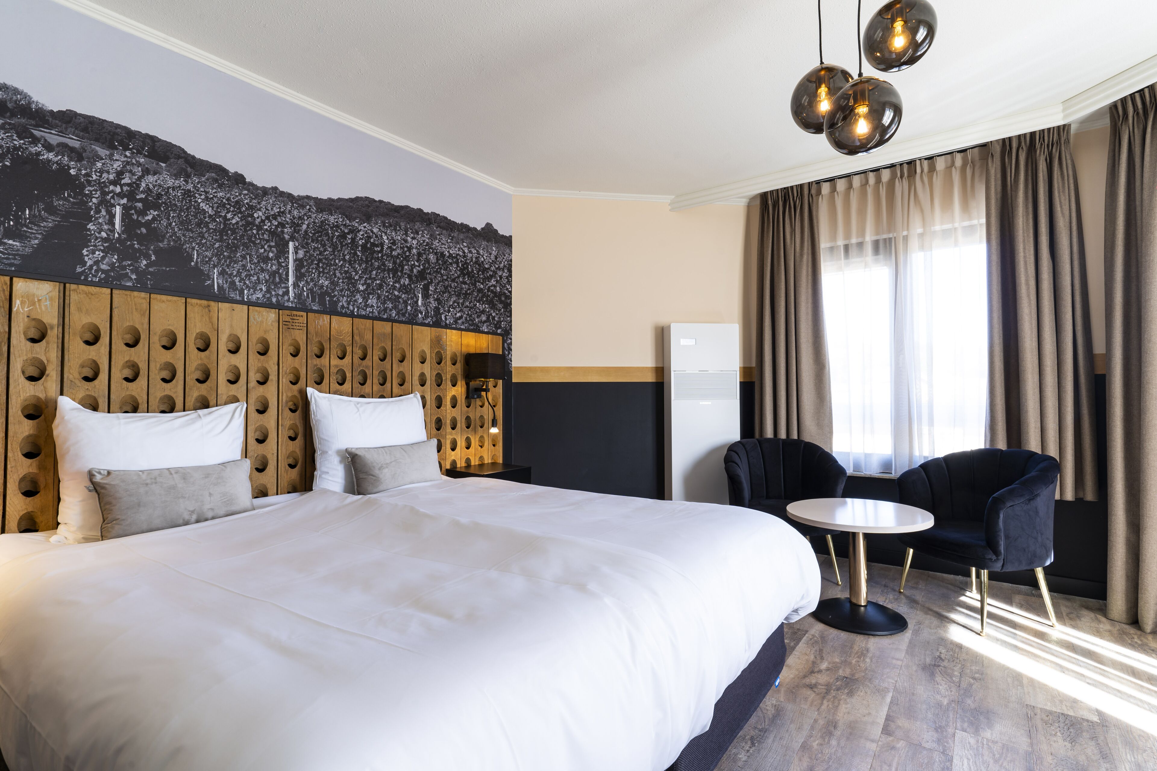 Een hotelkamer met een groot bed, een houten hoofdeinde, een ronde tafel, een zwarte stoel en een wandlamp.