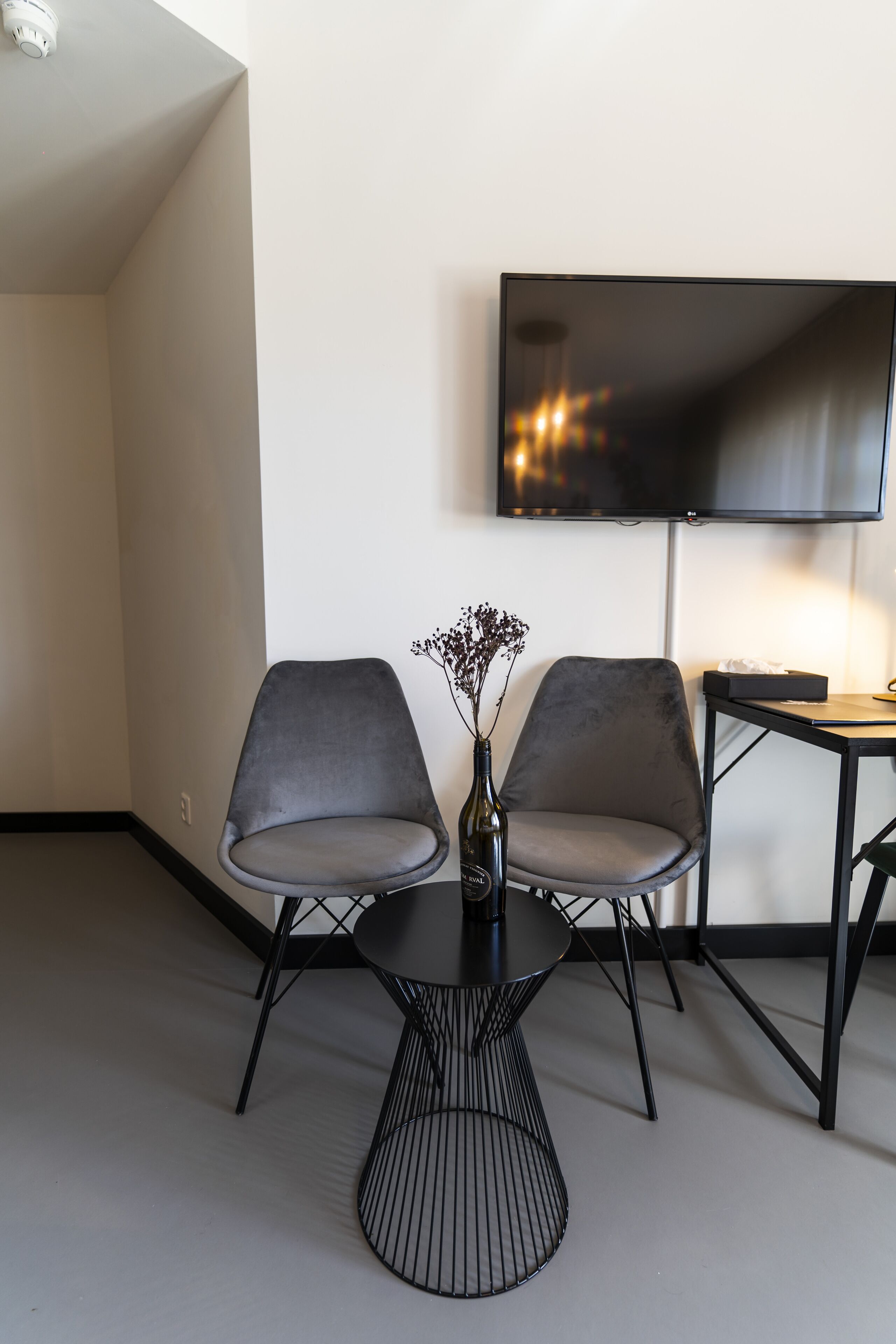 A modern room with two grey chairs, a small table, a vase with flowers, and a flat-screen TV mounted on the wall.