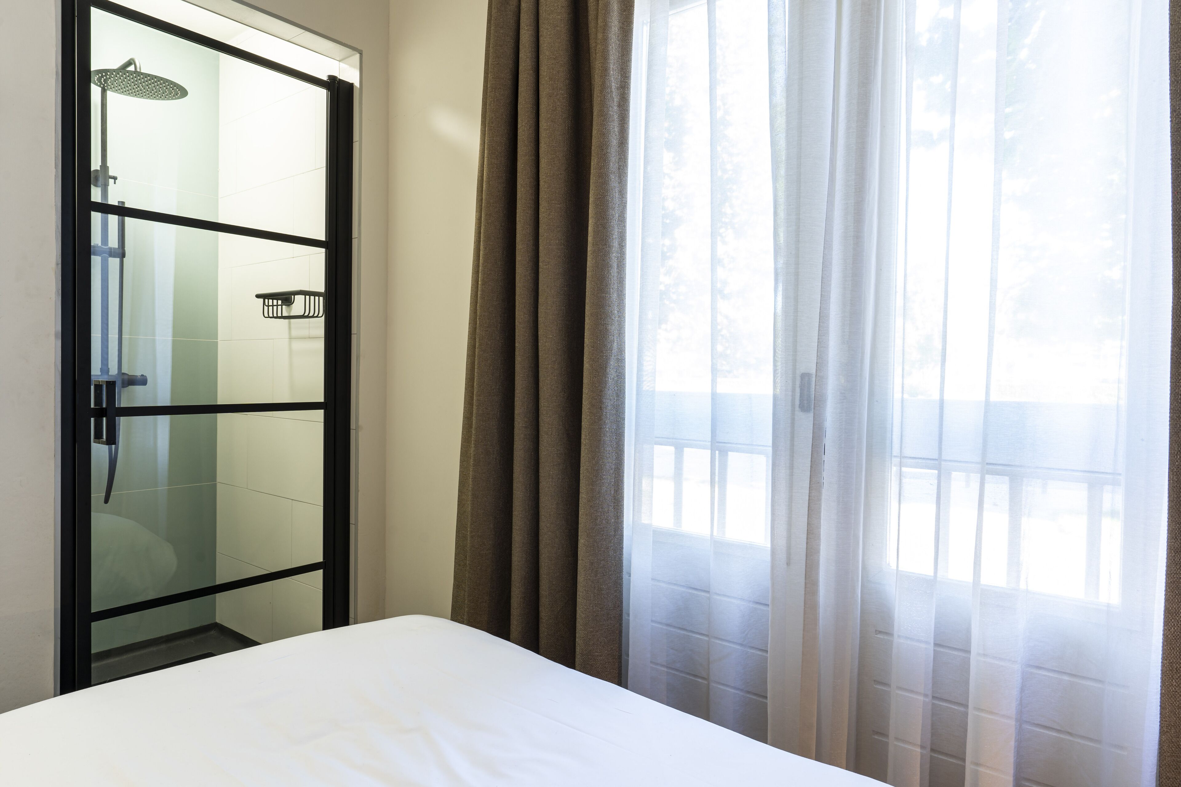 A hotel room with a bed, a glass shower enclosure, and a view of the outdoors through sheer curtains.