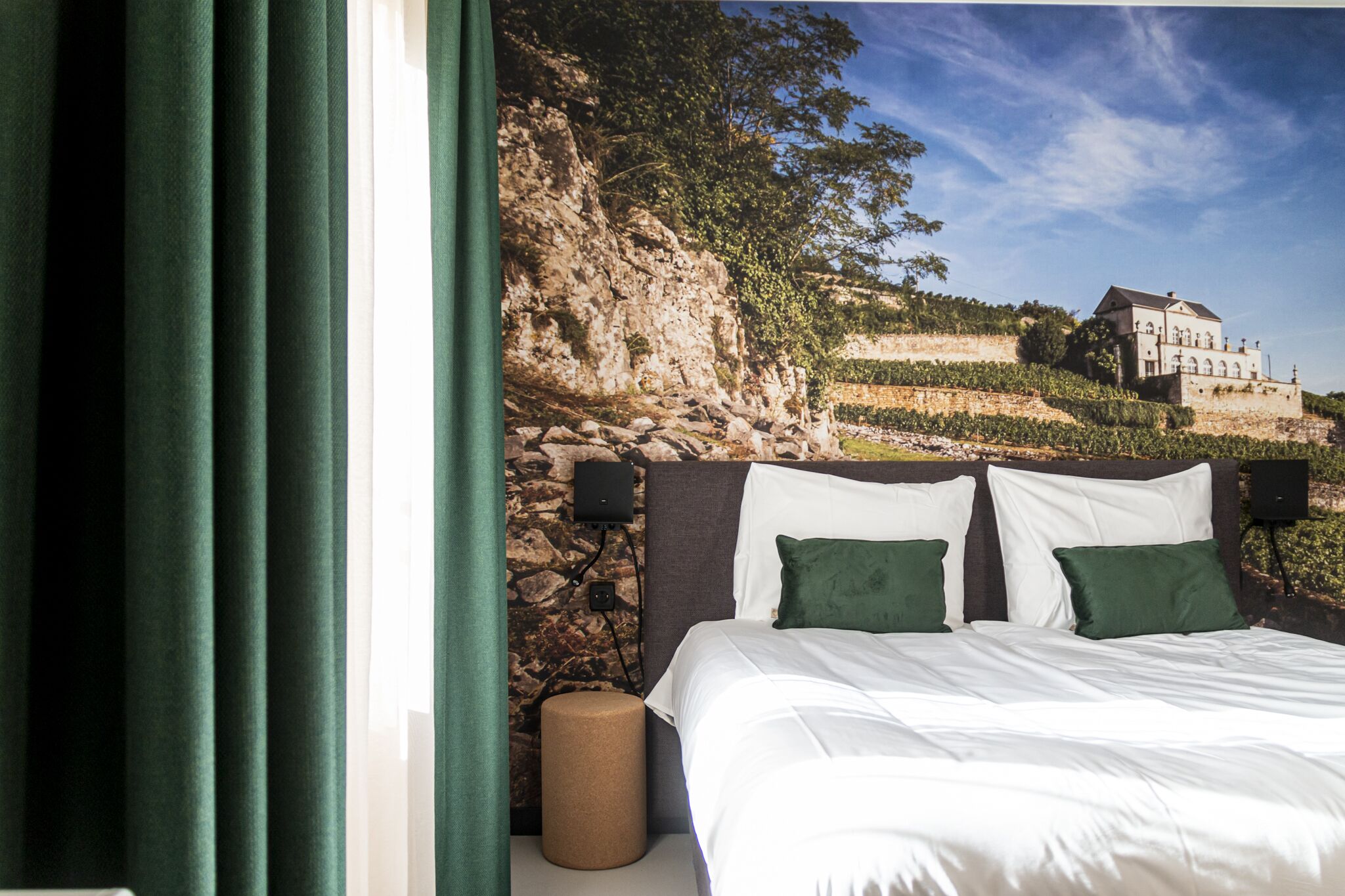 Een slaapkamer met een stenen muur, een bed met witte beddengoed en groene kussens, en uitzicht op een gebouw op een heuvel.